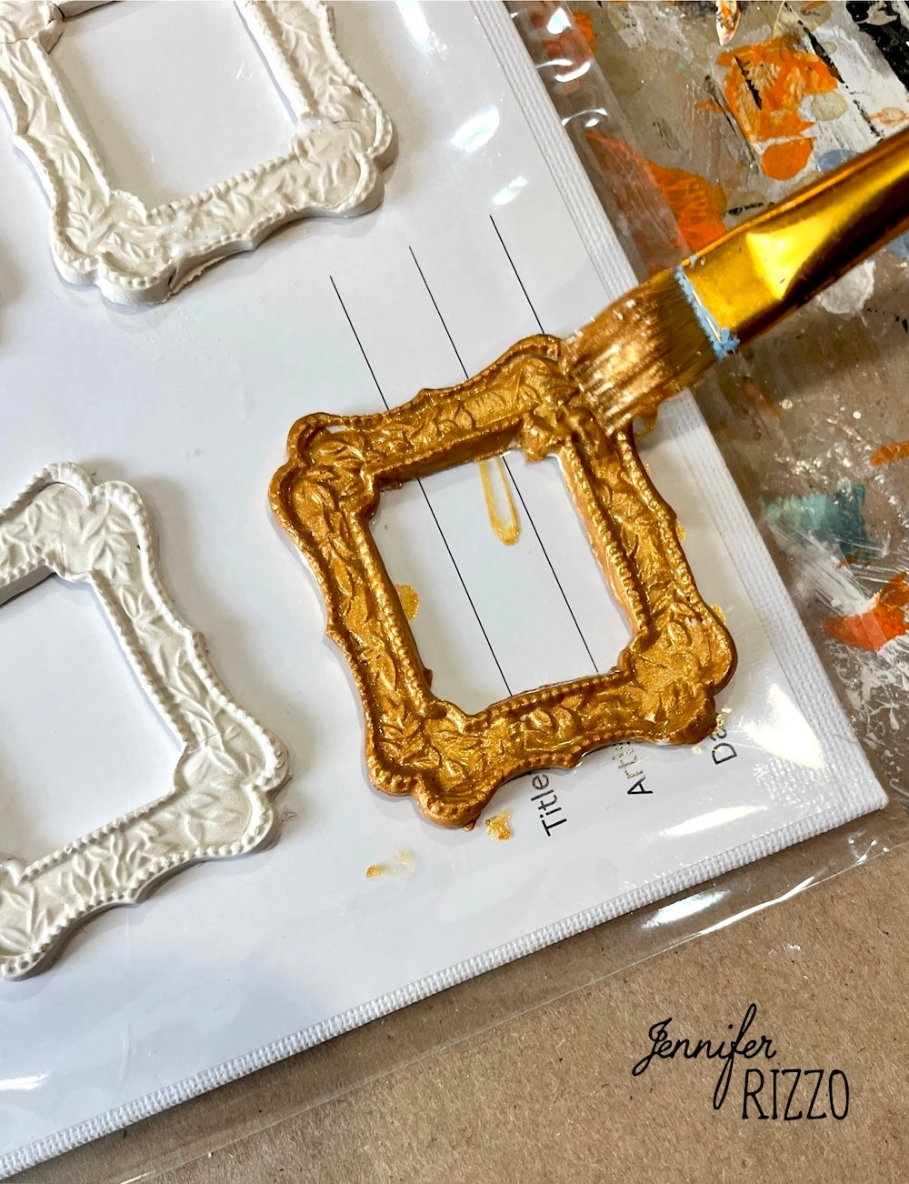 A hand uses a paintbrush to apply gold paint to a small, decorative picture frame; unpainted white frames are visible beside it on a plastic sheet.