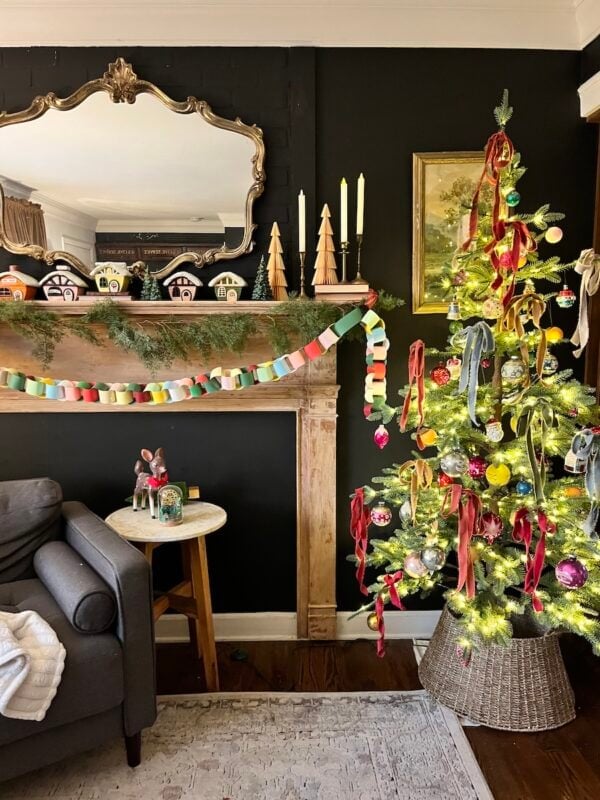 A decorated Christmas tree with colorful ribbons stands beside a fireplace mantel adorned with garland, candles, wooden trees, and small house ornaments in a cozy living room.