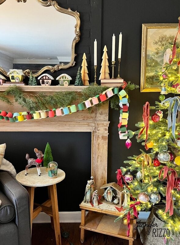 A decorated fireplace mantel with paper garland, small houses, wooden trees, and candles sets a nostalgic and vintage Christmas decorating tone. Nearby, a Christmas tree and nativity scene add to the charming holiday décor.