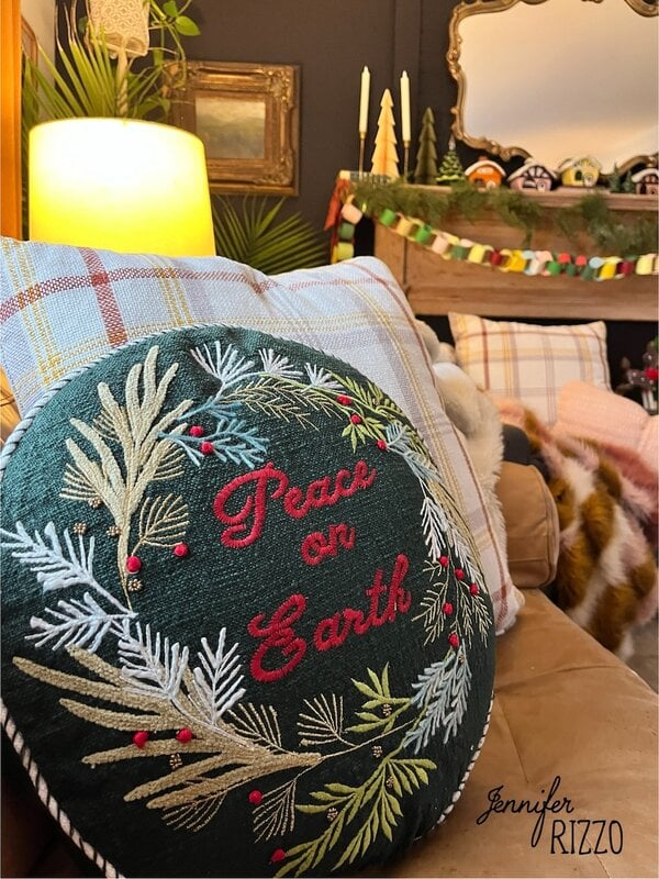 A round pillow with “Peace on Earth” and a wreath design sits on a tan sofa, adding a touch of nostalgic and vintage Christmas decorating to a warmly decorated living room filled with holiday cheer.