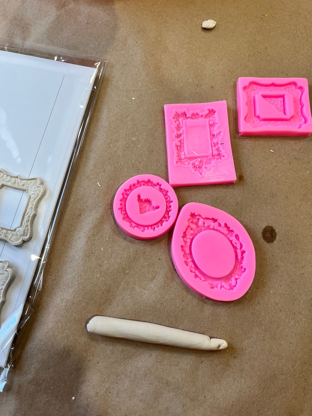 Pink silicone molds and white clay frame shapes are arranged on a brown surface, with a rolled piece of clay nearby.
