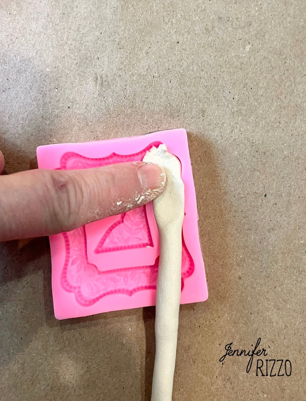 A hand presses a rolled piece of white clay into a pink silicone mold on a brown surface. "Jennifer Rizzo" is printed in the lower right corner.
