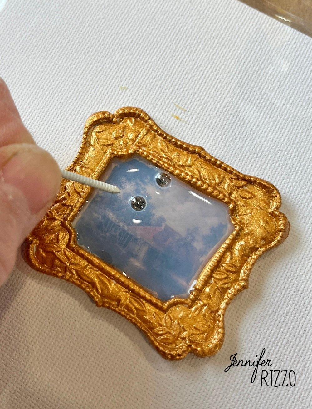 A hand uses a cotton swab to place resin and attach two small googly eyes onto a miniature framed artwork with a gold ornate border.
