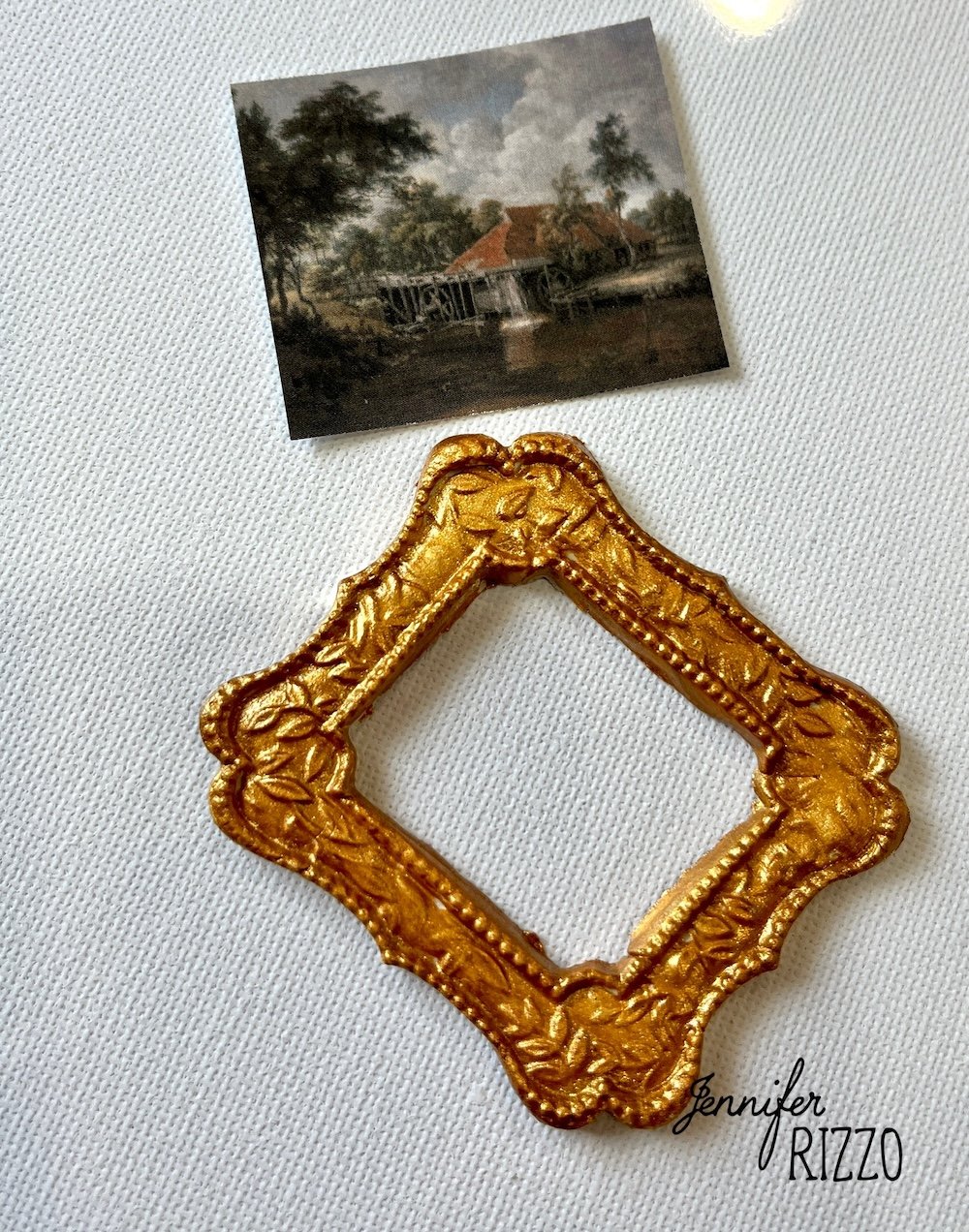 A small, ornate gold picture frame and a miniature landscape painting rest on a white textured surface; “Jennifer Rizzo” is written in the corner.