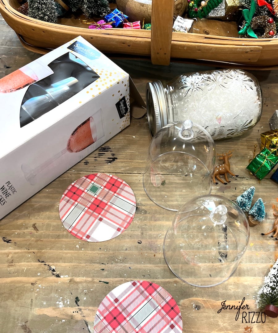 Two glass domes, plaid coasters, a box labeled "Plastic Wine Glasses," a snow-filled jar, and mini holiday decorations on a wooden table. Mini Snow Globe Cloches from Plastic Wine Glasses