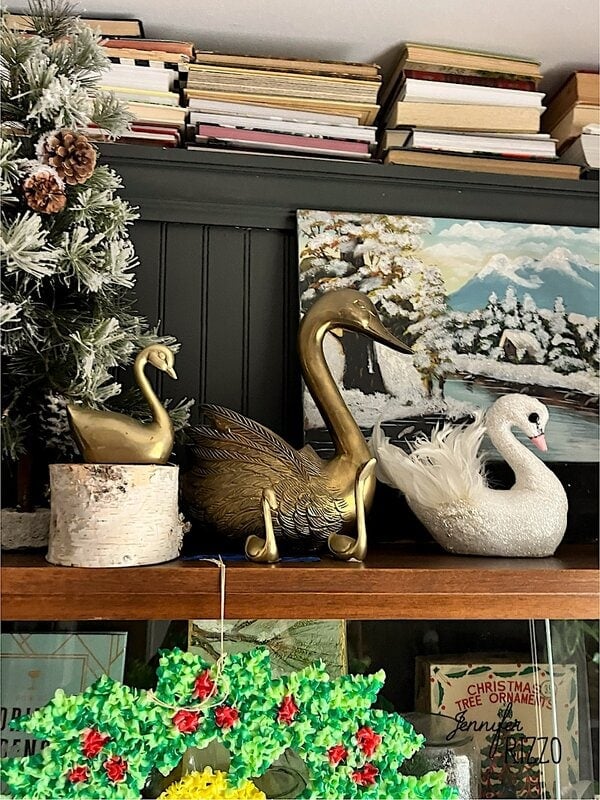 Three decorative swans—gold, brass, and glittery white—are displayed on a shelf with a snowy painting, frosted tree, stacked books, and holiday decor for a touch of nostalgic and vintage Christmas decorating charm.