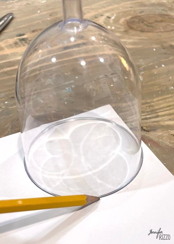 A pencil traces the rim of an upside-down wine glass onto a sheet of white paper on a wooden surface.