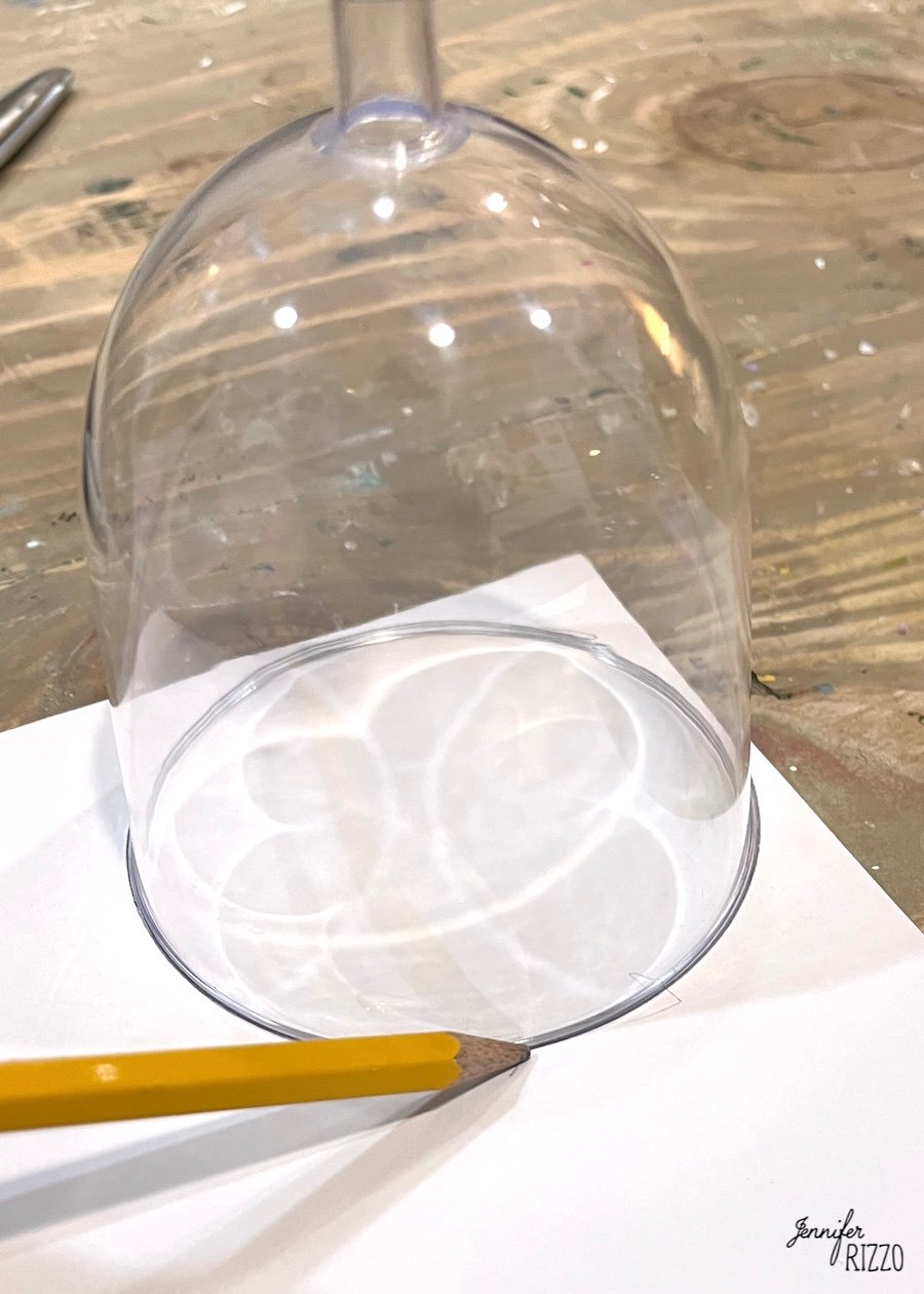 A pencil traces the rim of an upside-down wine glass onto a sheet of white paper on a wooden surface.