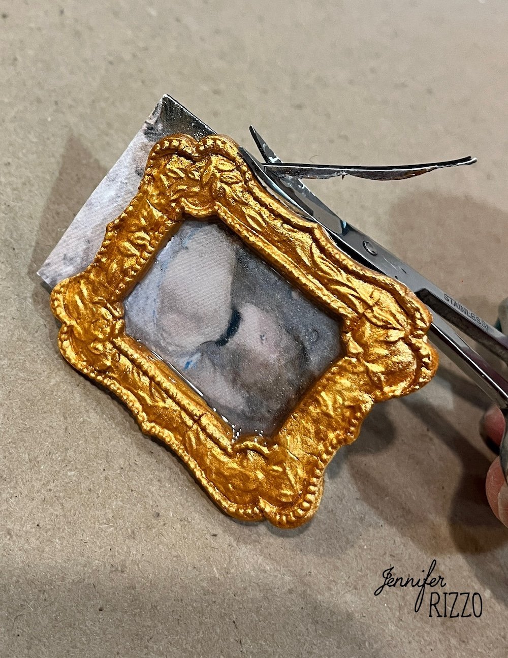 A hand cuts out a small printed photo with scissors, fitting it into an ornate, gold miniature picture frame on a beige surface.