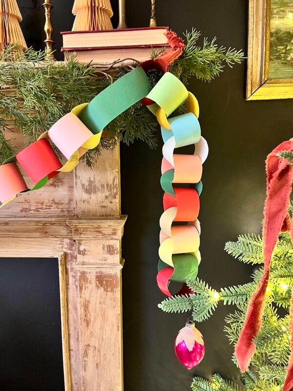 A colorful paper chain garland hangs from a mantel decorated with greenery, with a red ornament attached to the end near a lit Christmas tree.