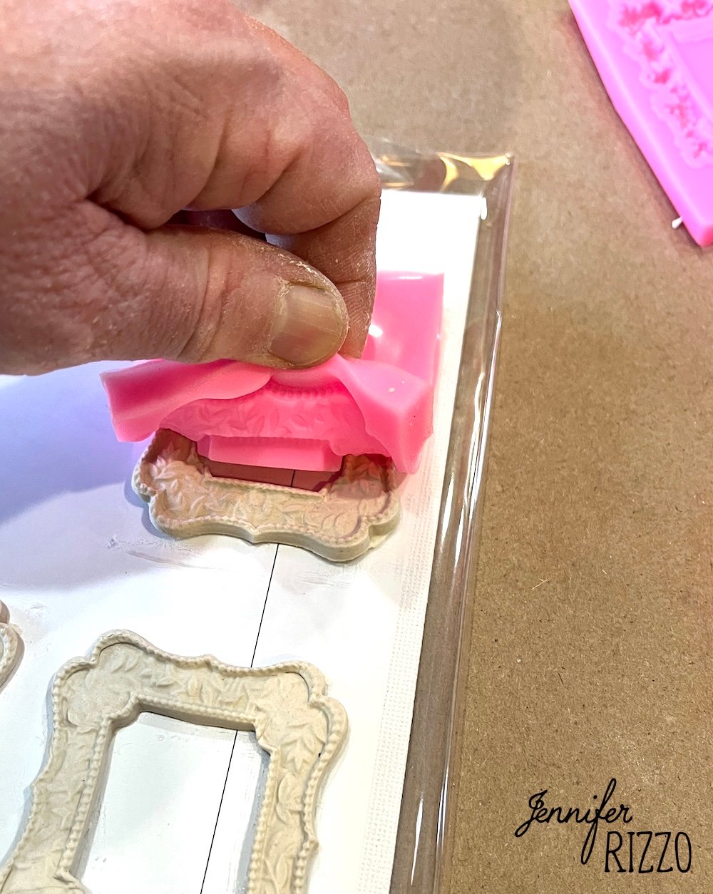 A hand presses a flexible pink mold over a decorative frame-shaped piece on a table, with other similar frame pieces nearby. The name "Jennifer Rizzo" appears in the corner.