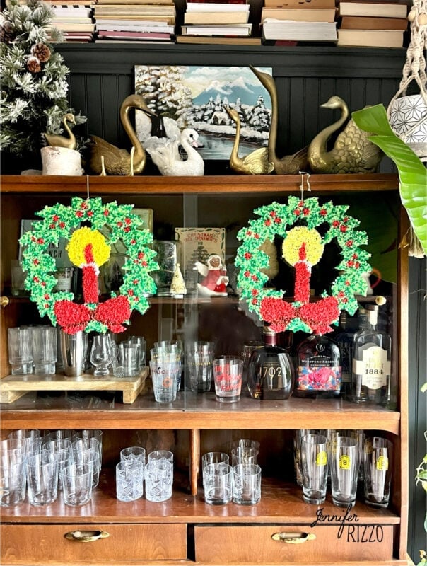 Two faux stained glass wreath decorations with candle designs evoke nostalgic and vintage Christmas decorating, hanging on a glass cabinet filled with various glassware, with swan figurines and books on top.
