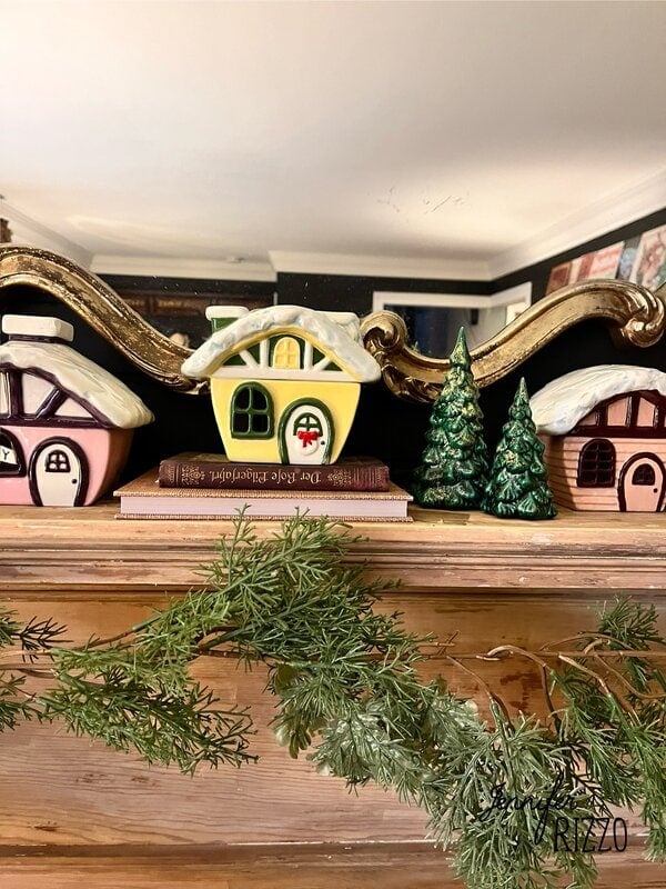 Small ceramic houses with snow-covered roofs evoke nostalgic and vintage Christmas decorating, displayed on a wooden mantle flanked by faux greenery and a book, with a decorative mirror in the background.
