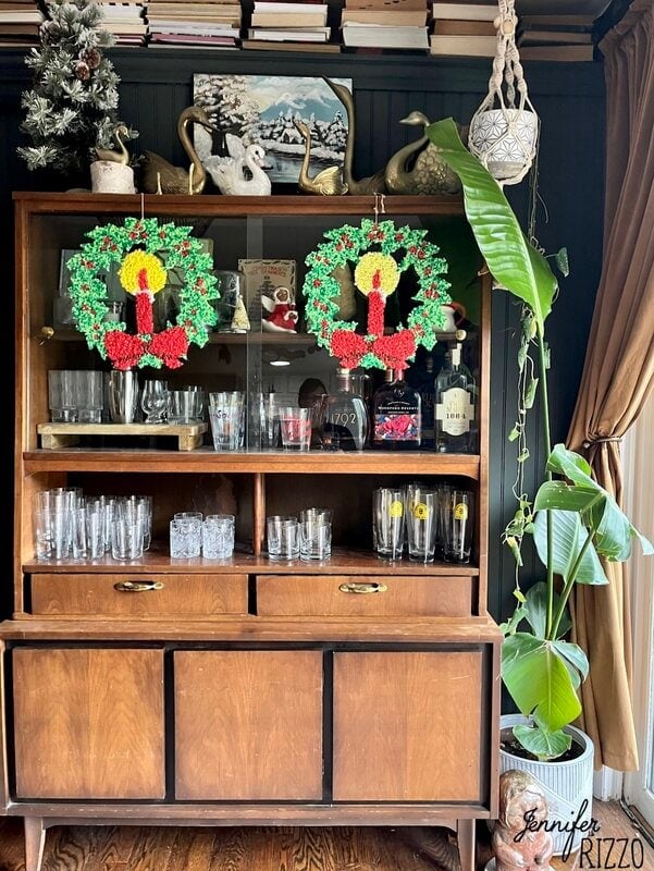 A wooden hutch with glassware, liquor bottles, and two crocheted Christmas wreaths hanging on the top doors creates a nostalgic and vintage Christmas decorating vibe. Plants and books are on and beside the hutch.