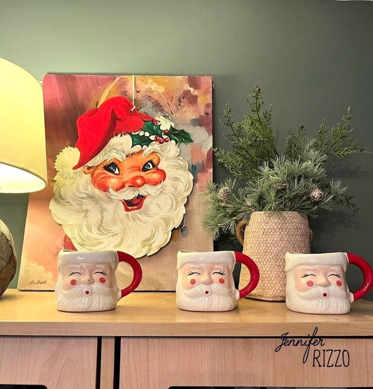 Three Santa Claus mugs are displayed on a light wooden surface in front of a Santa painting, capturing the charm of nostalgic and vintage Christmas decorating. A potted pine plant and a lamp complete this festive scene.