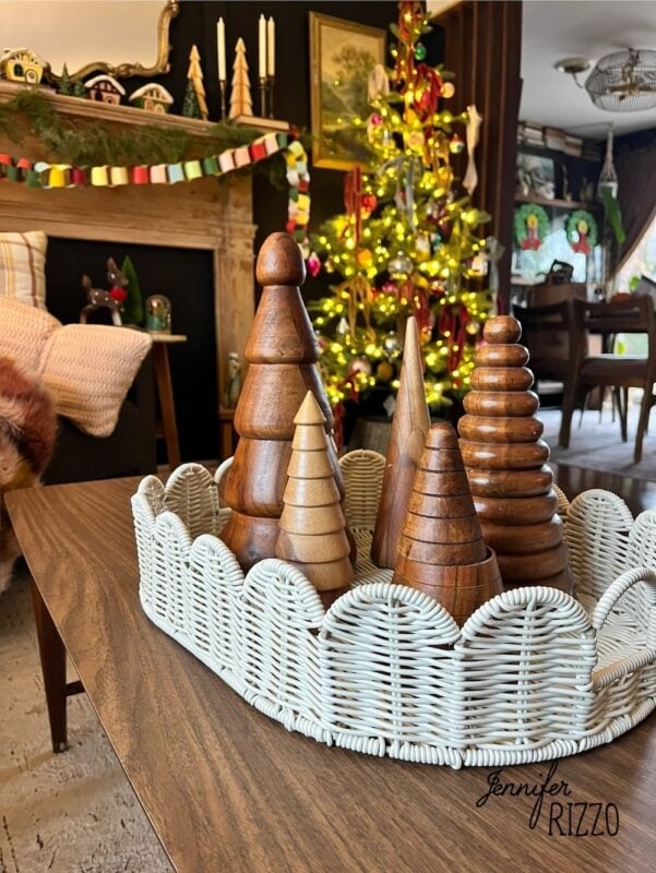 A wicker tray with wooden trees sits on a coffee table in a living room decorated for Christmas, capturing the charm of nostalgic and vintage Christmas decorating with a lit tree and paper garland in the background.