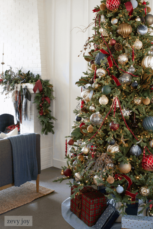A decorated Christmas tree with ornaments and lights stands next to wrapped gifts; a fireplace with a garland is visible in the background.