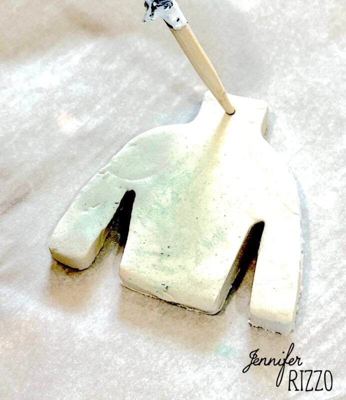 A wooden stick is making a hole in a sweater-shaped piece of white clay, placed on a sheet of parchment paper.