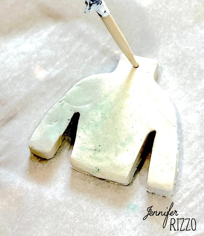 A wooden stick is making a hole in a sweater-shaped piece of white clay, placed on a sheet of parchment paper.