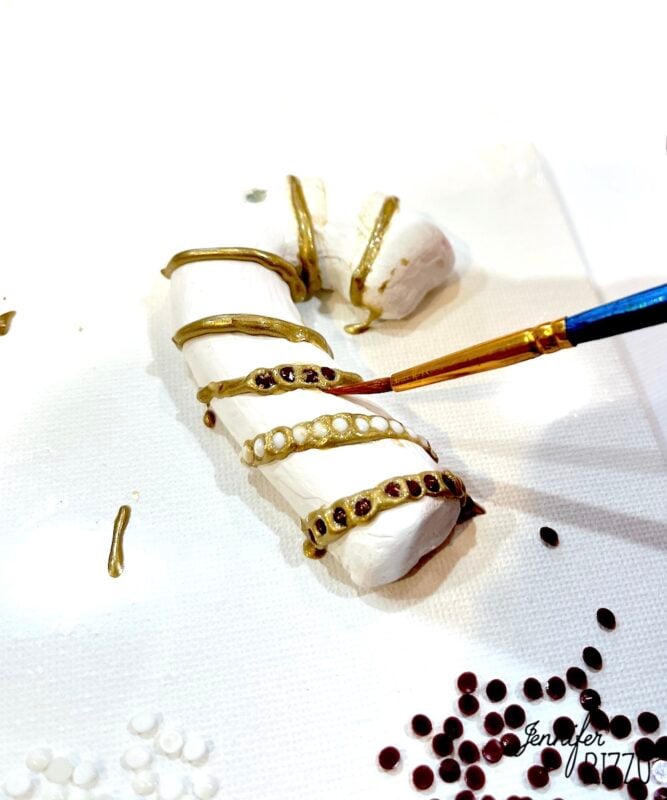 A hand paints gold lines and places beads on a white candy cane-shaped object on a white surface, with scattered red and white beads nearby.