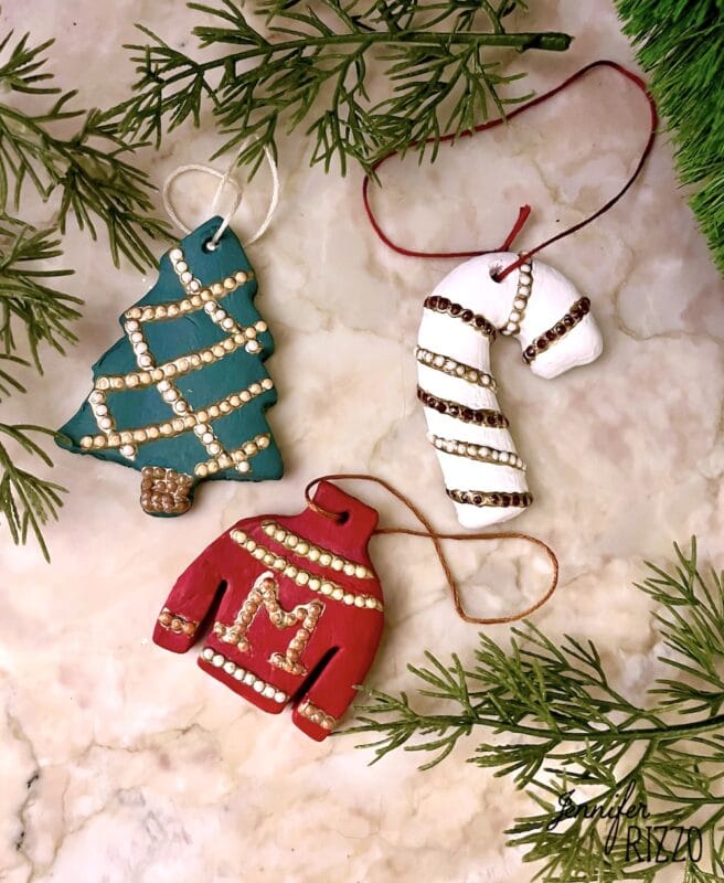 Three handmade holiday ornaments—a green tree, a red sweater with an "M," and a white candy cane—are decorated with beads and placed on a surface with evergreen branches.