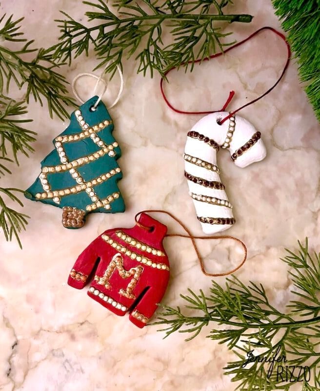 Three handmade holiday ornaments—a green tree, a red sweater with an "M," and a white candy cane—are decorated with beads and placed on a surface with evergreen branches.