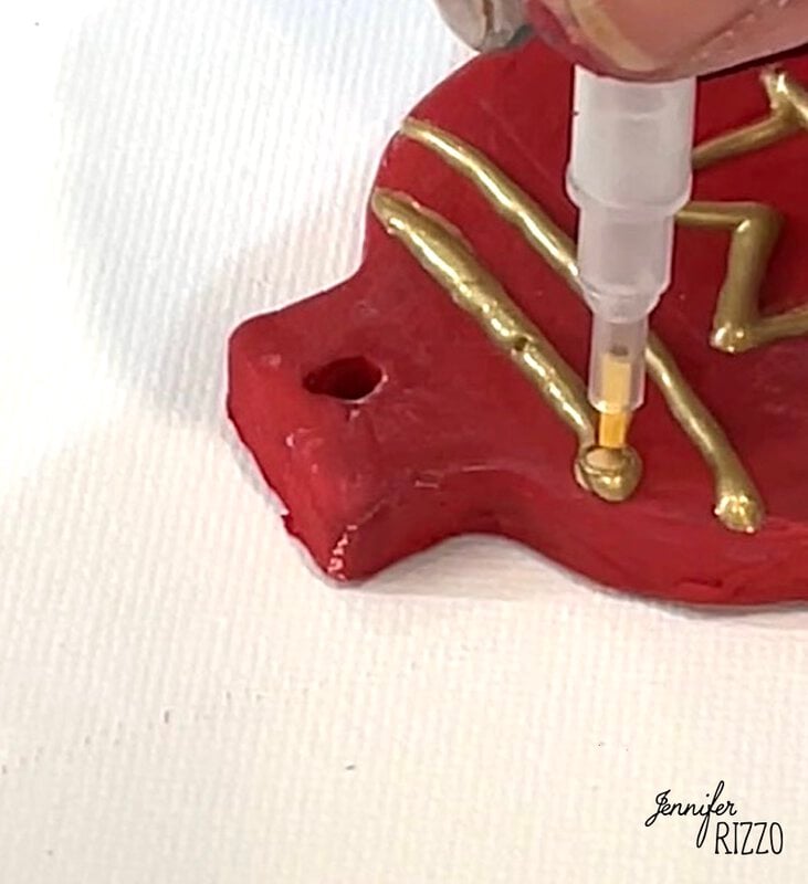 A hand uses a gold paint pen to decorate a red clay ornament with gold lines and dots on a white surface.