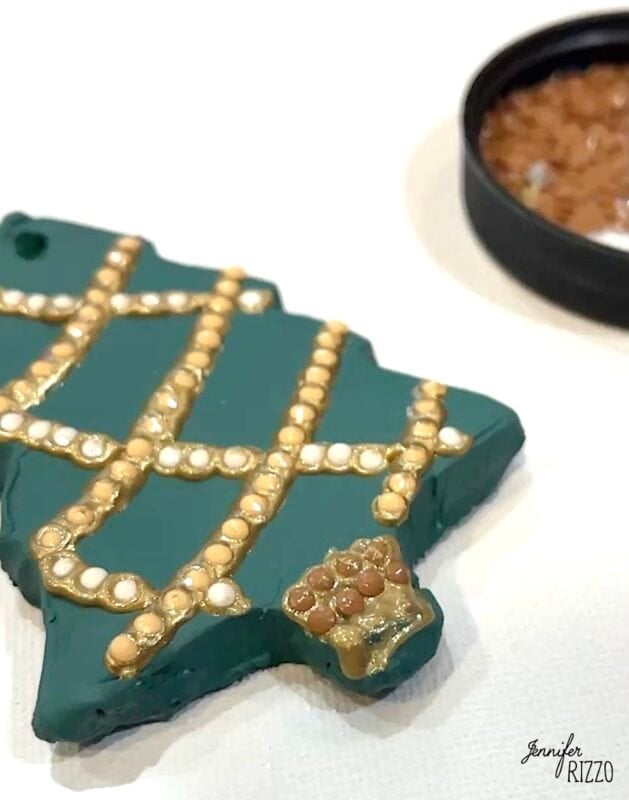 A green Christmas tree ornament with gold and white dot decorations lies on a white surface; small brown beads are nearby.