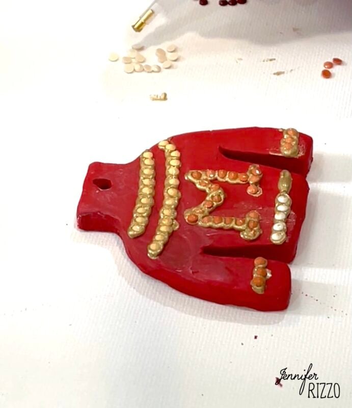 A red sweater-shaped ornament decorated with gold and white beads lies on a white surface with scattered beads nearby.