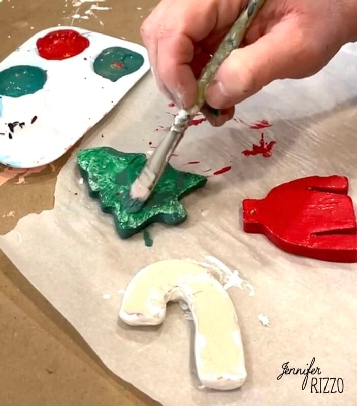 A hand paints a green Christmas tree-shaped ornament with a brush; red sweater and unpainted candy cane ornaments are nearby on parchment paper.