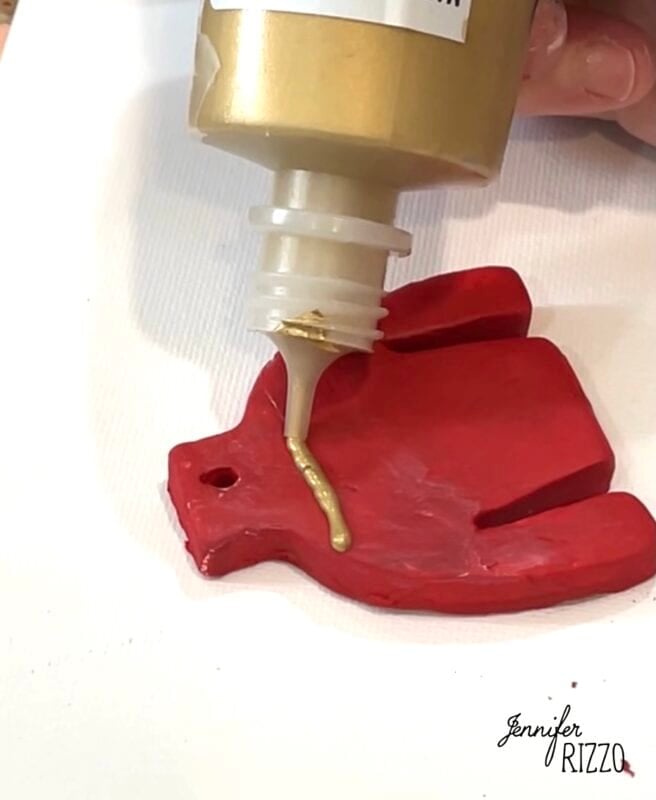 A hand-shaped red clay ornament is being decorated with a line of gold paint squeezed from a bottle.