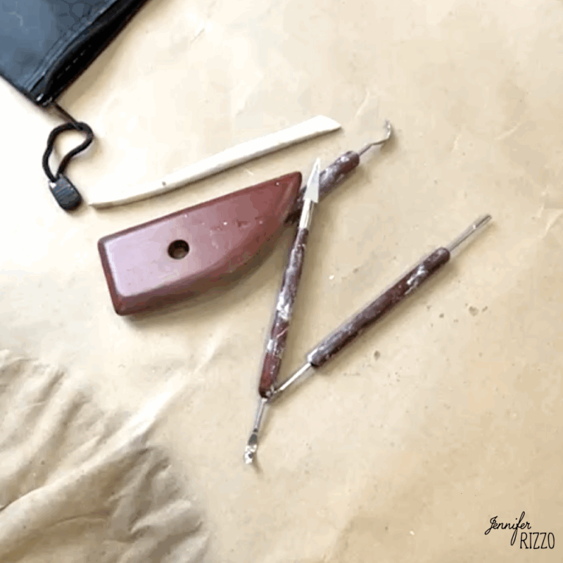 Four pottery sculpting tools and a black pouch are arranged on a brown surface, with some clay residue visible on the tools.