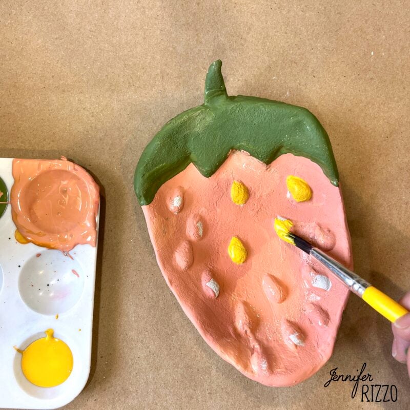 A hand uses a paintbrush to paint yellow spots on a strawberry-shaped clay dish; a palette with pink and yellow paint is beside it.