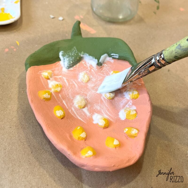 A brush applies white glaze to a ceramic piece shaped and painted like a strawberry, resting on a brown surface.