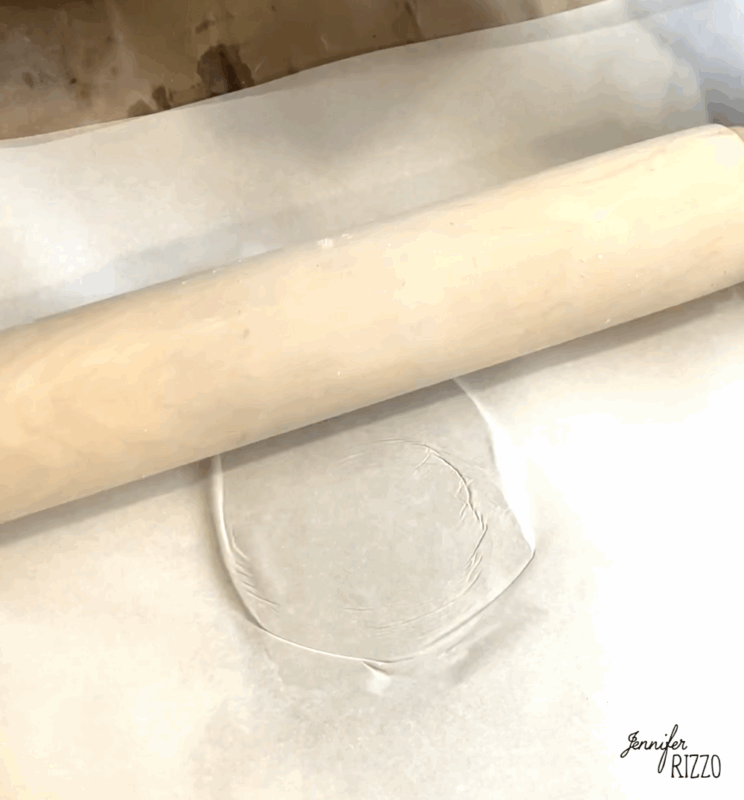 A rolling pin flattening a piece of clear material between two sheets of parchment paper on a light-colored surface.