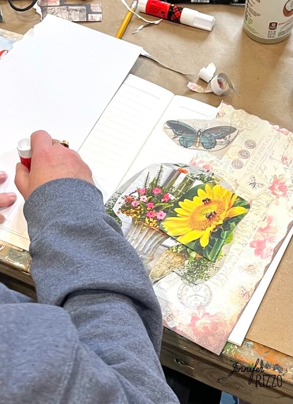 A person gluing a piece of decorative floral paper into a notebook; the paper features a butterfly and a vase with sunflowers and pink flowers.