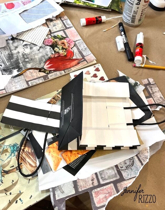 A workspace with cut paper, a striped paper bag, glue sticks, and various craft supplies arranged on a brown surface.