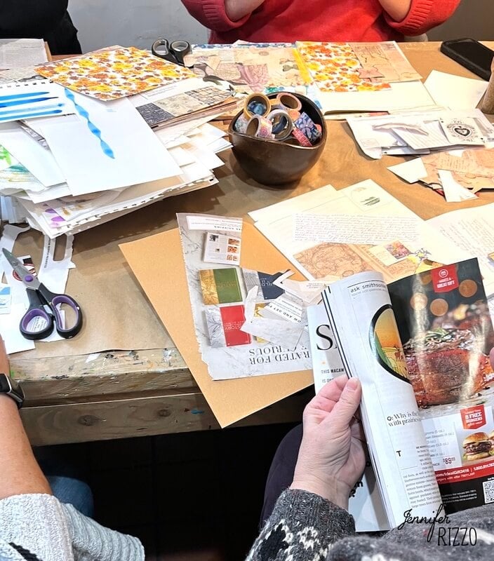 Three people sit at a table covered with scrapbooking supplies, papers, scissors, and magazines, engaging in a crafting activity.