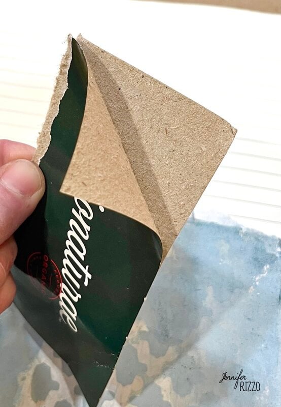 A hand is peeling a green adhesive label from a piece of brown cardboard.