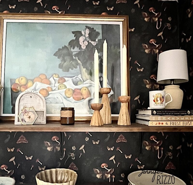 A shelf with a framed still life painting, two tall candles in wooden holders, a small lamp, stacked books, a mug, a candle, and a decorative clock against a dark floral wallpaper.