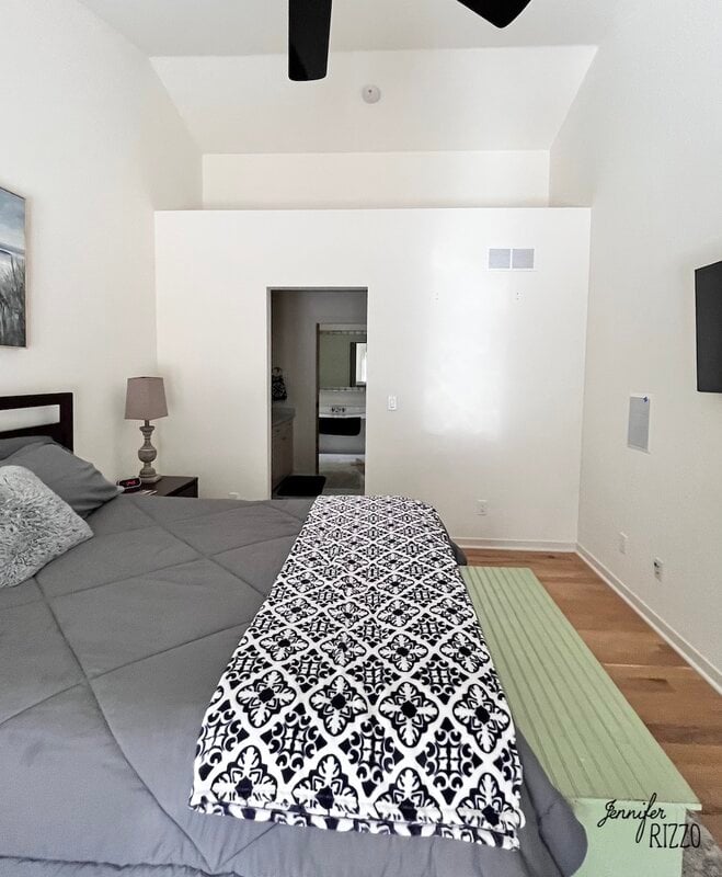A modern bedroom with a gray bedspread, patterned blanket, side table with lamp, wall-mounted TV, and a doorway leading to another room.