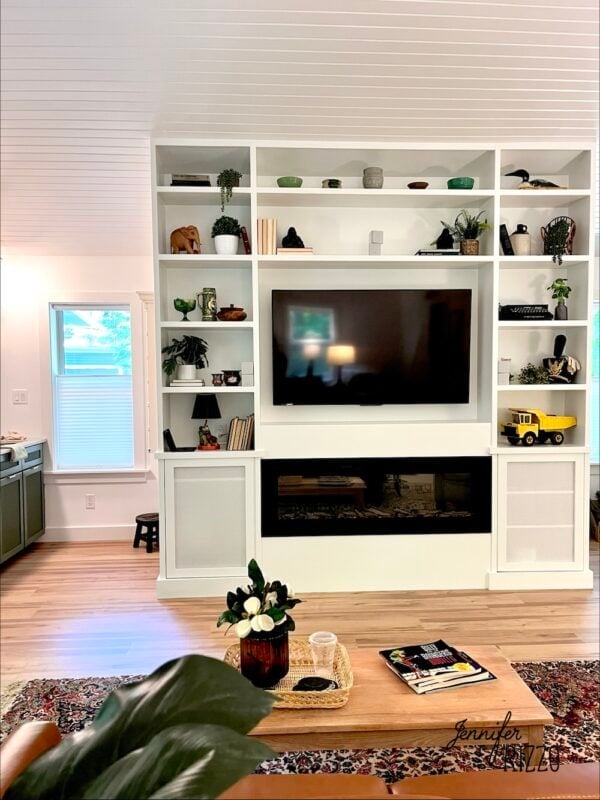 A modern living room with built-in white shelves, a wall-mounted TV, decorative items, a yellow toy truck, a fireplace, and a wooden coffee table with plants and magazines.