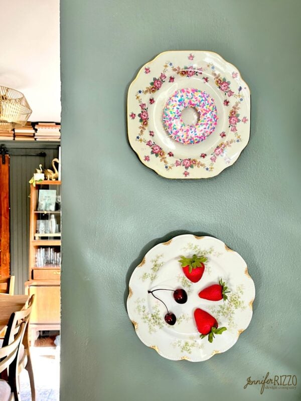 Two decorative plates hang on a green wall; the top plate has a donut with sprinkles, while the bottom plate holds three strawberries and two cherries.