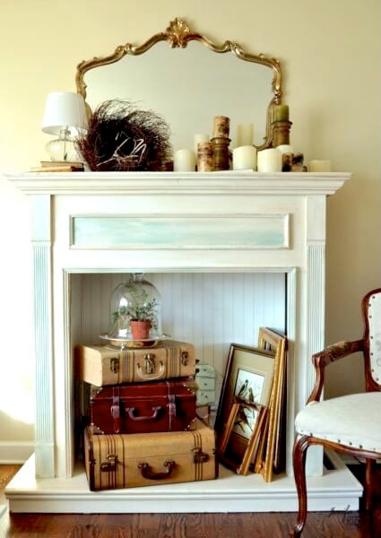 A decorative fireplace mantel displays candles, a nest, and a lamp. Inside the fireplace are stacked vintage suitcases, framed art, and a glass cloche with a potted plant.