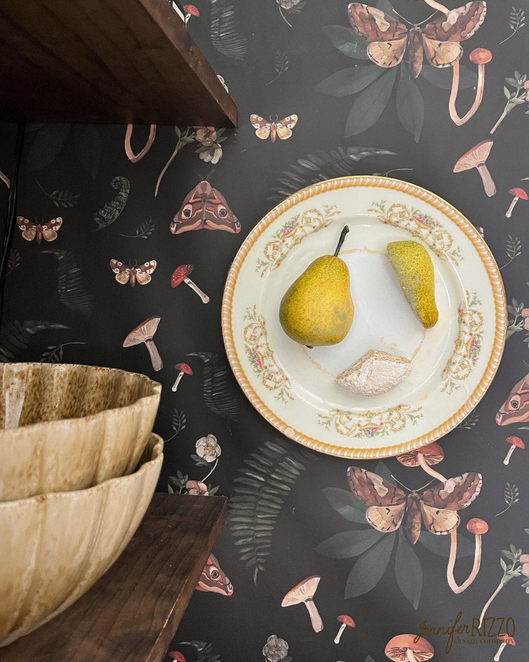 A decorative plate with two pears and a pastry rests on a shelf against a dark, botanical wallpaper with mushroom and moth motifs. Wooden bowls are stacked nearby.