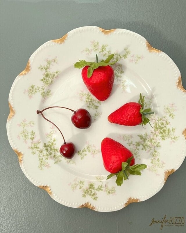 Three strawberries and two cherries on a floral-patterned vintage plate with a scalloped edge, set on a gray surface.