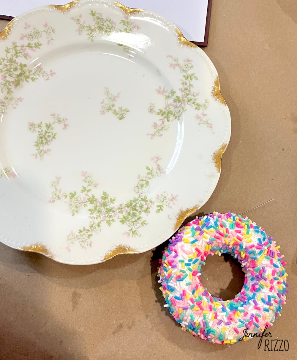 A decorative floral plate with gold edges sits next to a donut covered in colorful sprinkles on a brown surface.