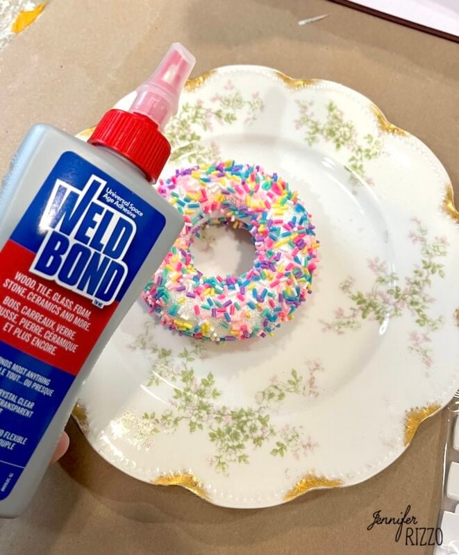 A hand holds a bottle of Weldbond glue near a faux donut covered in colorful sprinkles, placed on a decorative plate.