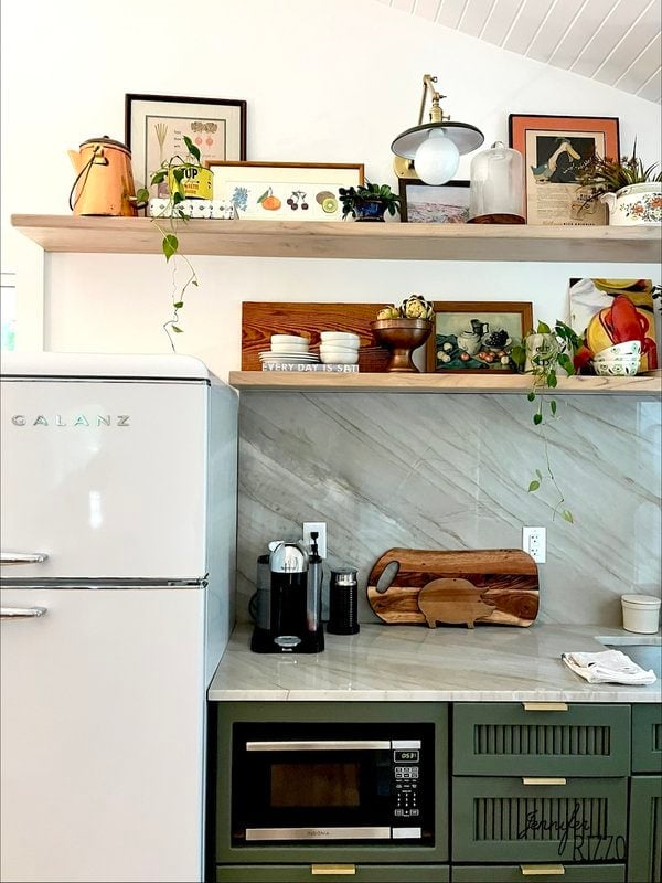 A kitchen corner with a white fridge, coffee maker, wooden cutting boards, green cabinets, marble backsplash, and open shelves with dishes, plants, and framed art.