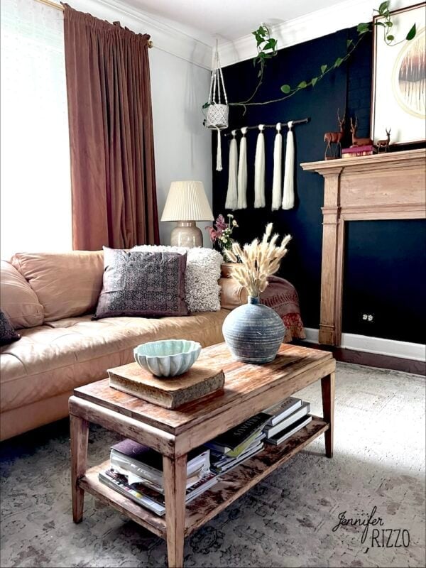 A cozy living room with a tan leather sofa, wooden coffee table with books and decor, dark accent wall, and pampas grass in a vase.
